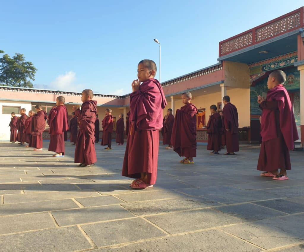 Eine Gruppe junger Mönche in roten Roben steht draußen im Hof eines Klosters und betet in einer Reihe bei sonnigem Wetter.