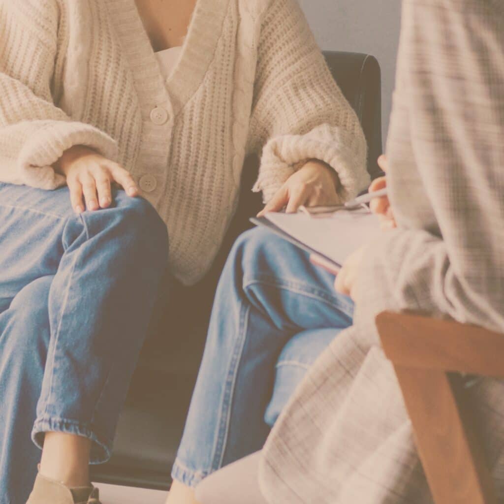 Nahaufnahme einer psychologischen Beraterin, die Notizen auf einem Klemmbrett macht, während sie mit einer Person in einem Strickpullover spricht.